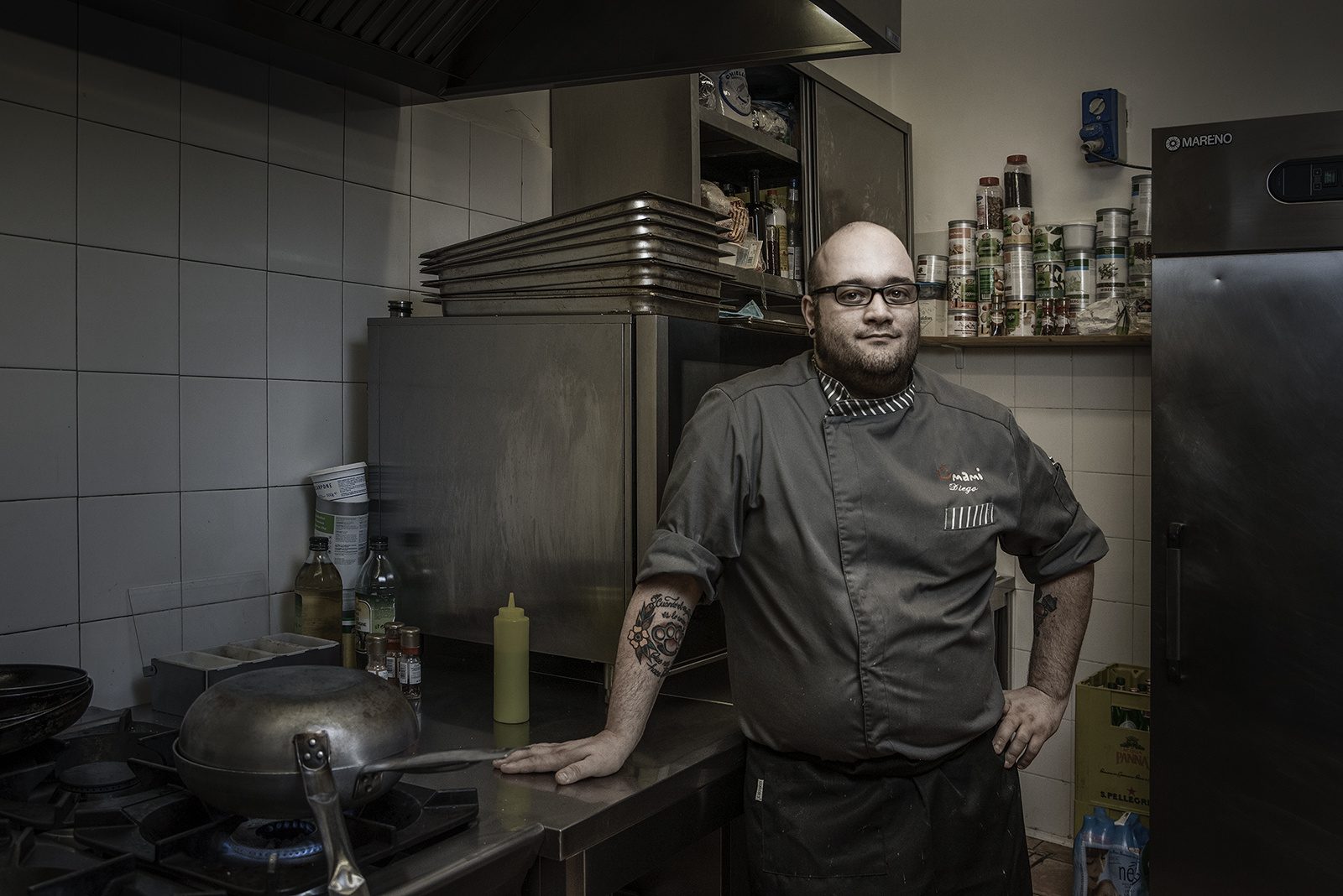 Uno chef con la giacca scura appoggiato al piano della cucina del suo ristorante