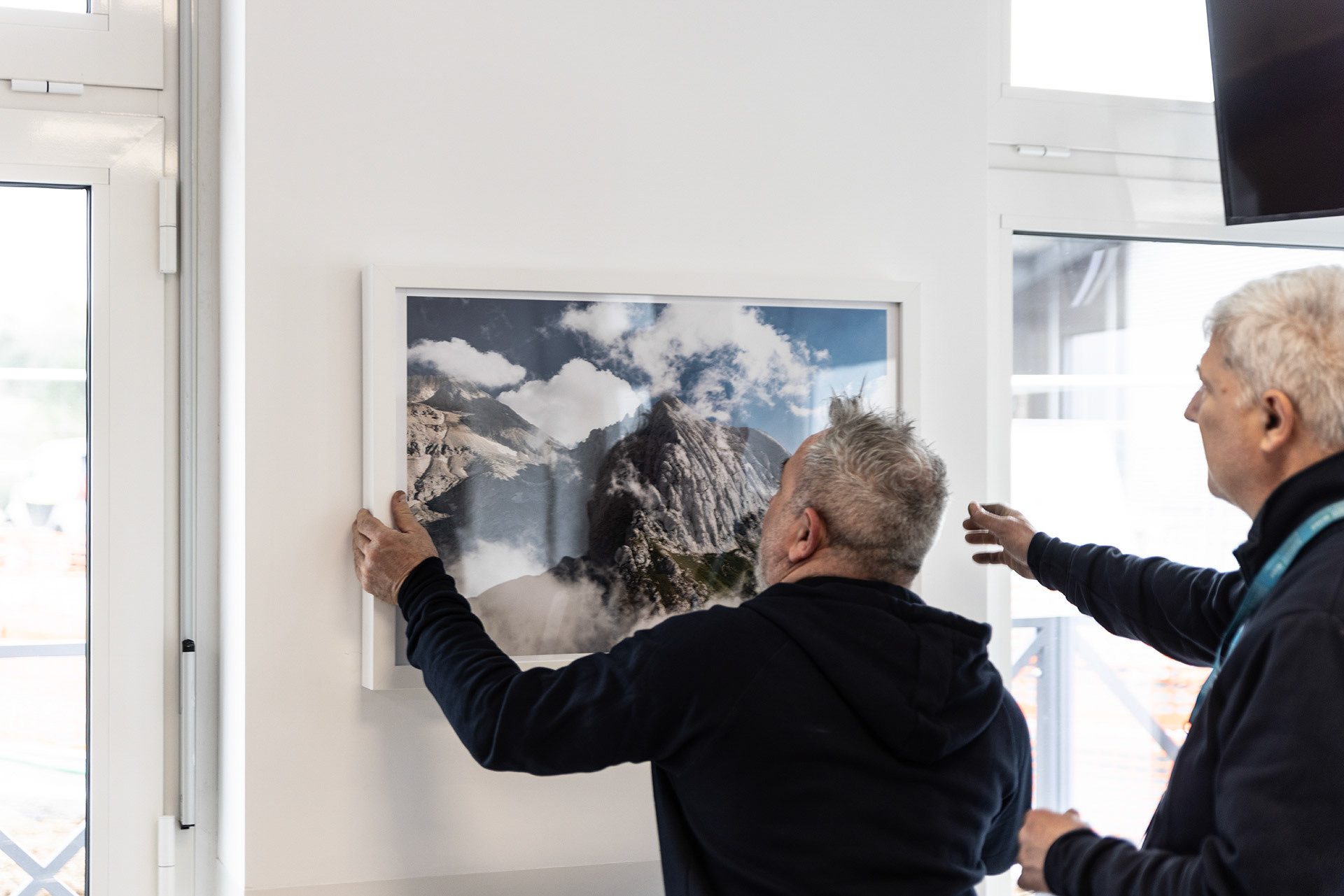 Due persone appendono una fotografia di montagna incorniciata nella sala d’attesa del Pronto Soccorso del Policlinico Gemelli