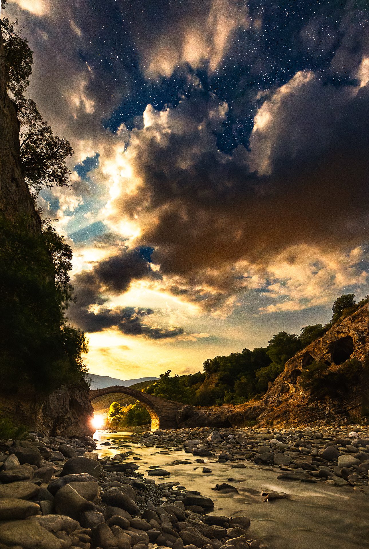 Un fiume tra le rocce che scorre sotto un antico ponte ad arco, con un cielo di nuvole al tramonto, Albania