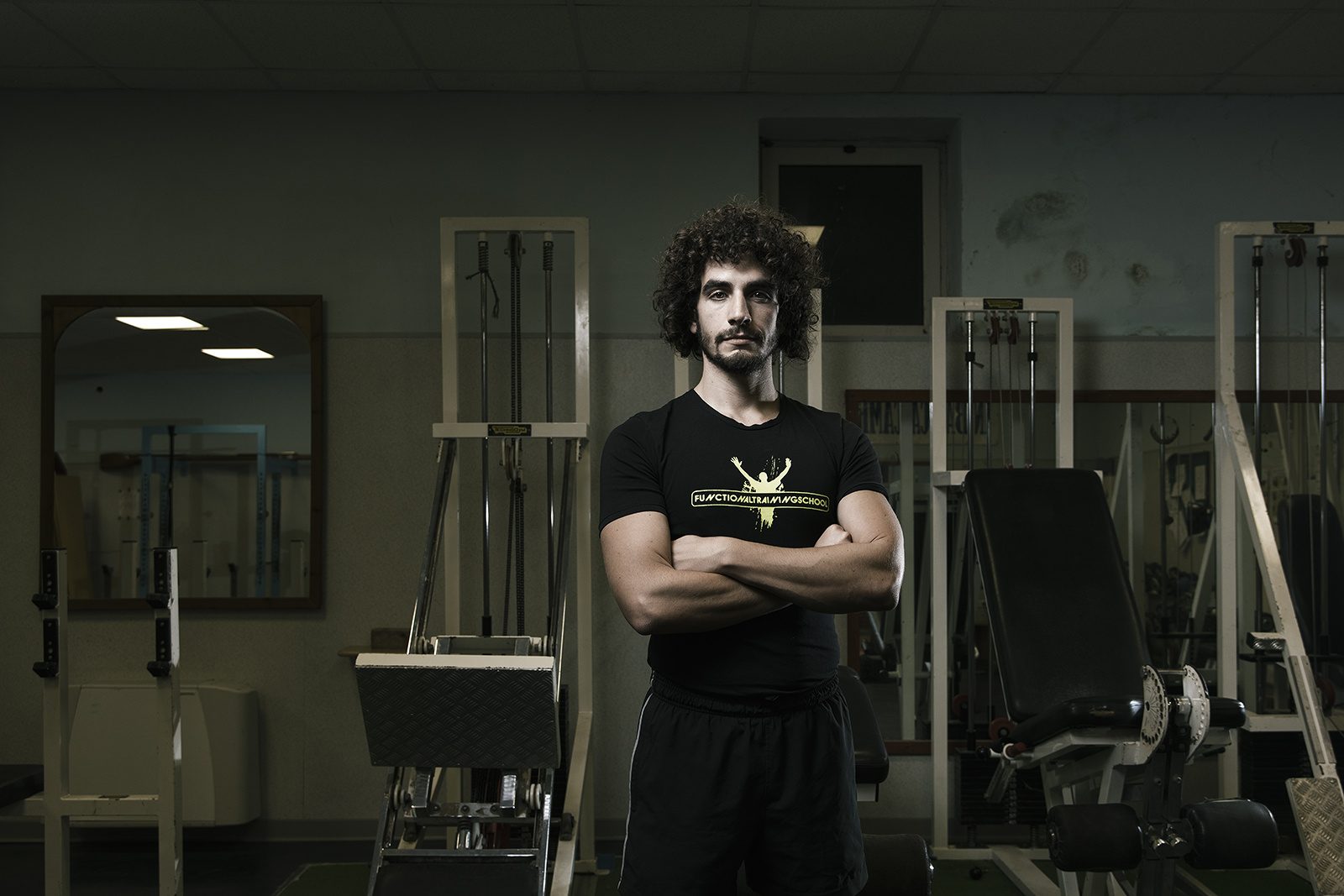 Un giovane uomo con i capelli ricci, a braccia conserte tra le macchine di una palestra