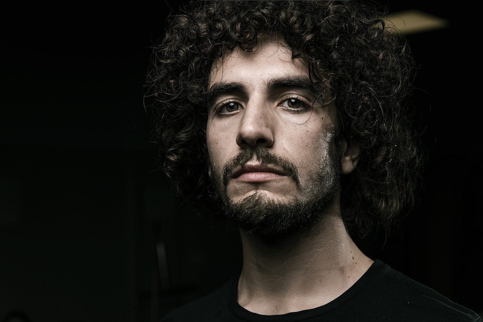 Primo piano di un giovane uomo con i capelli ricci e la barba, su fondo scuro