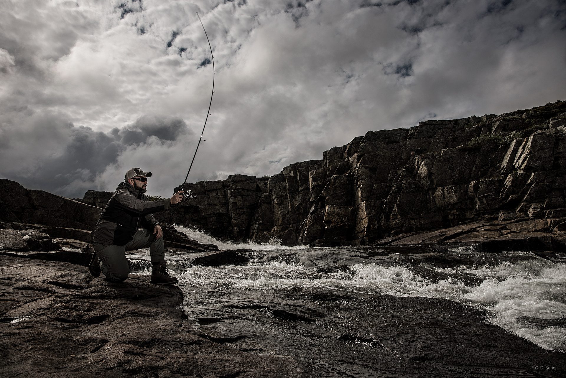 Pescatore inginocchiato su una roccia mentre lancia con la canna da spinning, accanto a una rapida sotto pareti di roccia e un cielo carico di nuvole, Norvegia
