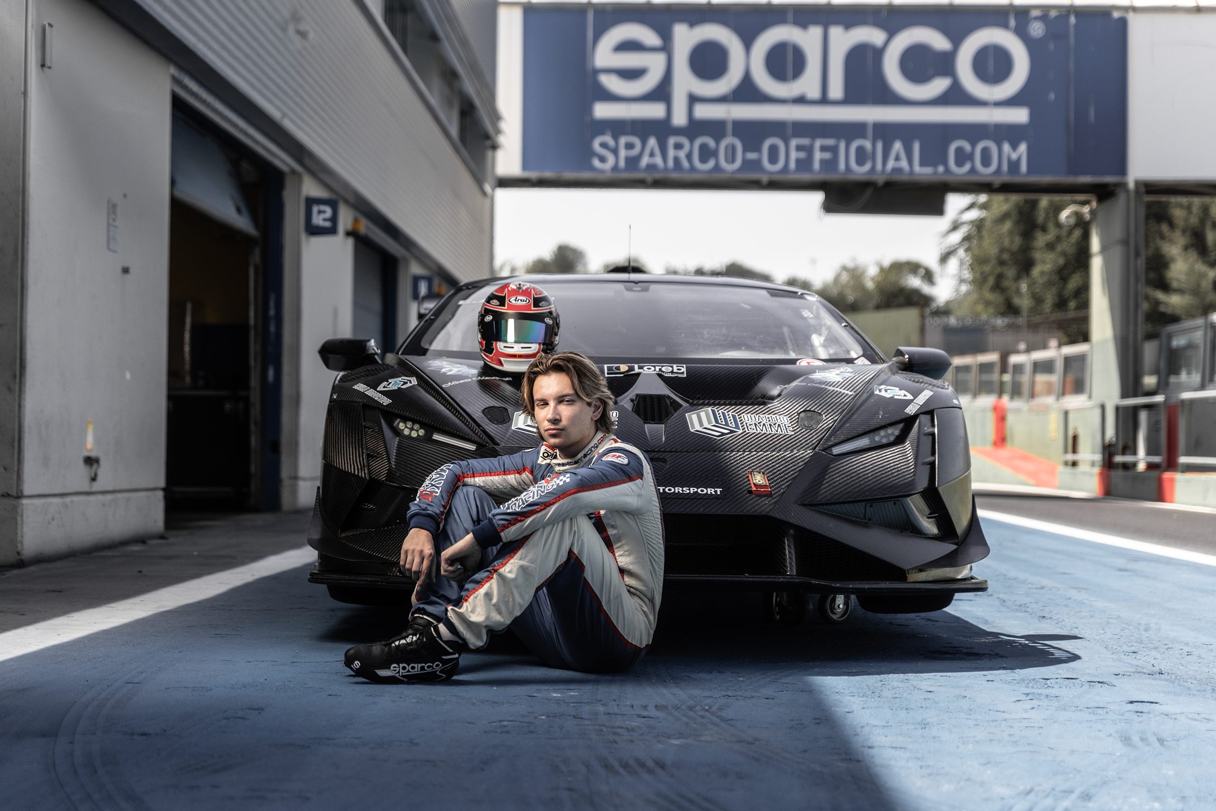 Pilota seduto davanti a un’auto GT nel box, sotto l’insegna Sparco