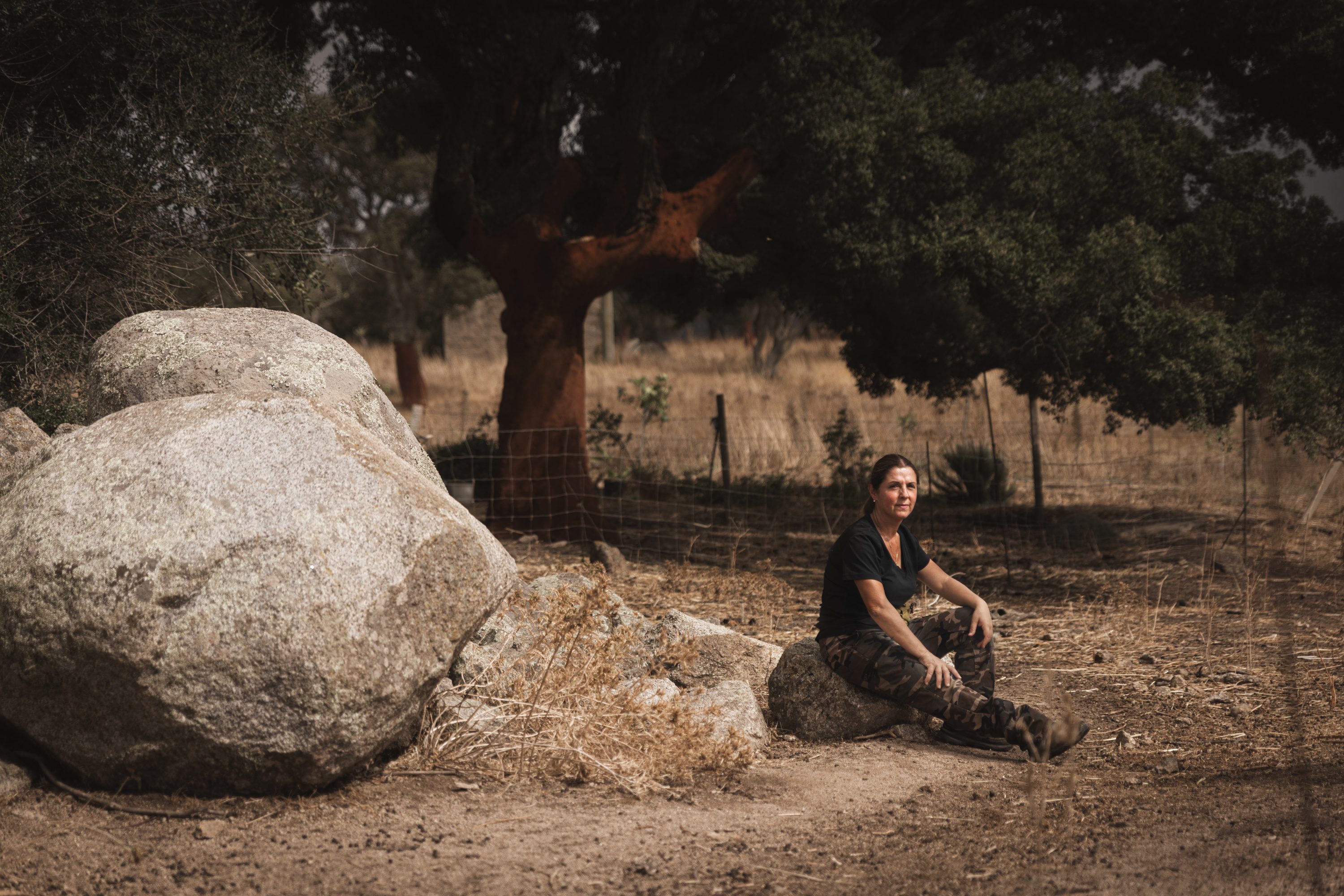Una donna seduta a terra all’ombra di una sughera, accanto a grandi massi di granito